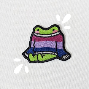 May include: A green frog wearing a purple, blue, and white striped sweater. The frog is smiling and has a happy expression.
