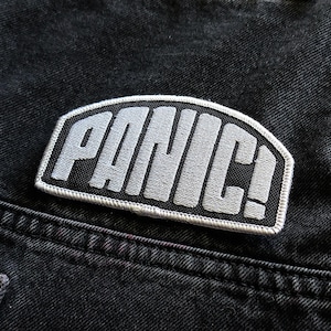 May include: A black and white embroidered patch with the word "PANIC!" in white letters on a black background. The patch is sewn onto a black denim jacket.