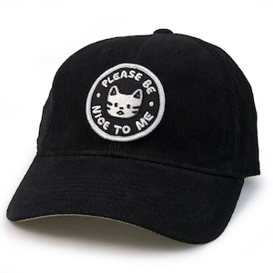 Może przedstawiać: Czarna bejsbolówka z kordurowej tkaniny z białym haftowanym naszywką z napisem "Proszę, bądź miły dla mnie" i przedstawiającą kreskówkową kotkę.
