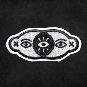 May include: White embroidered patch with a black eye in the center and two white eyes on either side. The eyes have black pupils and white irises. The patch has a white outline and a black background.