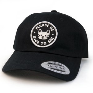 May include: Black baseball cap with a white embroidered patch that says "Please Be Nice To Me" and features a cartoon cat. The cap is adjustable.