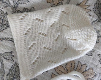 Könnte beinhalten: Eine cremefarbene Strickhaube mit einem zarten Lochmuster auf dem Hauptteil und einem strukturierten kraus rechts gestrickten Rand an der Vorderseite. Die Haube verfügt über zwei lange, passende Strickbänder, die an der Basis befestigt sind.