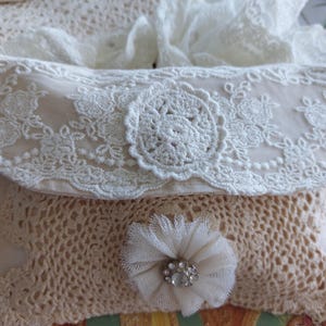 May include: A cream-coloured fabric pouch featuring an intricate crochet lace overlay. The top flap is adorned with a circular lace medallion, while the front body displays a gathered tulle flower centred with a small, clear, faceted crystal accent.