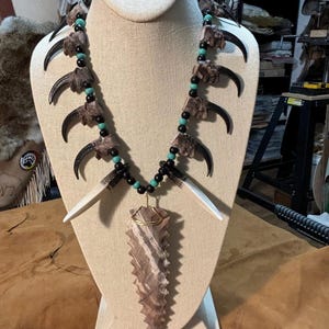 Puede incluir: Un collar hecho de garras de oso, cuentas turquesa y cuentas negras con un colgante de punta de flecha marrón y blanco.