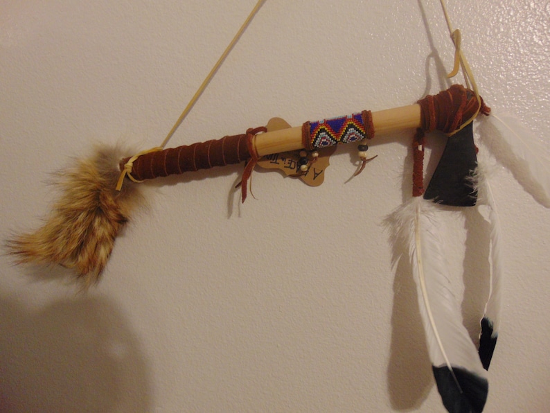 May include: A wooden peace pipe with a brown leather wrap, beaded accents, and a white and black feather. The pipe is hanging on a white wall with a brown tag that says "Alice-M-Art".