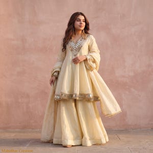 Könnte beinhalten: Ein cremefarbenes dreiteiliges Outfit bestehend aus einer kurzärmeligen Tunika, einer weiten Hose und einem passenden Dupatta. Die Tunika verfügt über einen V-Ausschnitt und einen Saum, die mit silberfarbenen Spiegeln und Stickereien verziert sind.