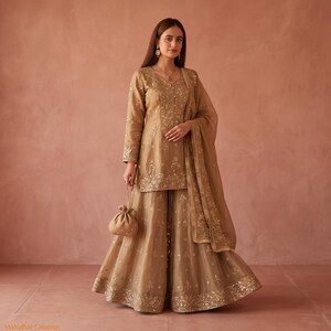 Peut inclure: Un ensemble trois pièces beige doré composé d'une tunique à manches longues avec broderies complexes, d'une jupe évasée assortie avec un large ourlet orné et d'une dupatta coordonnée. Une petite pochette à cordon est tenue à la main.