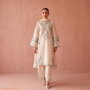 Könnte beinhalten: Ein cremefarbenes Set aus einer langärmeligen Tunika und einer passenden weiten Hose mit aufwendigen blauen Stickereien an Ausschnitt, Ärmelbündchen und Saum. Das Ensemble enthält einen passenden Schal mit blauen Quasten und den Text Mahidhar Creation.