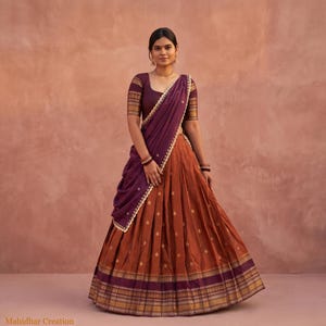 Puede incluir: Un conjunto de lehenga choli que presenta una blusa de color morado oscuro con mangas cortas y bordes dorados, combinada con una falda plisada de color naranja óxido adornada con pequeños motivos dorados y un dobladillo ancho morado y dorado. Un dupatta morado.