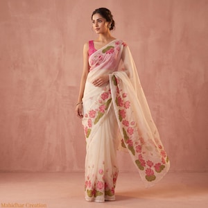 May include: A cream-colored saree featuring a vibrant pink lotus flower pattern with green leaves along the border and hem. The garment is paired with a sleeveless bright pink blouse. The text Mahidhar Creation is visible in the bottom left corner.