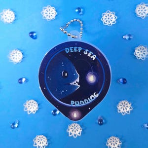 May include: A blue keychain with a deep sea creature design. The keychain has the words "DEEP SEA PUDDING" and a cartoon anglerfish illustration. The background is blue with decorative snowflake and teardrop accents.
