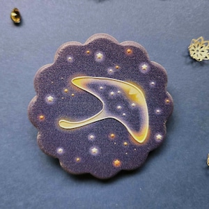 May include: A dark purple, scalloped-edge pin with a gold wishbone design and white and gold stars. The pin is surrounded by small gold and amber-colored beads and two gold flower-shaped embellishments on a dark blue surface.