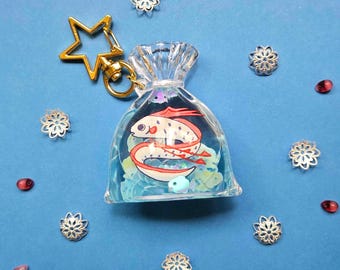 May include: A clear, fish-shaped keychain with a gold star-shaped clasp. The keychain contains a colorful illustration of a fish with red and white details. The background is a light blue with decorative silver snowflake charms and pink and red beads.