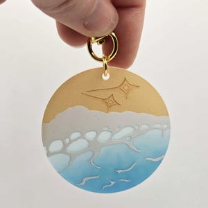 Puede incluir: Un llavero redondo con un diseño de escena de playa. La parte superior es de color beige, con estrellas grabadas. Debajo, un diseño de olas blancas y grises se transforma en un océano azul. Se adjunta un cierre dorado.