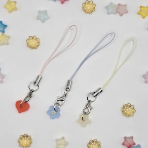 May include: Three pastel-colored phone charms with silver-tone clasps. One charm is a red heart, another is a blue flower, and the third is a yellow star. Scattered around are star and flower-shaped beads.