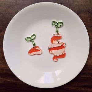 Puede incluir: Dos pendientes caprichosos sobre un plato blanco. Uno es una pequeña forma de serpiente naranja y blanca con un gancho verde en forma de hoja. El otro es una serpiente más grande envuelta alrededor de una manzana, adornada con flores.