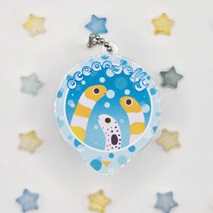 May include: A round, blue and white Ocean Jelly keychain with a silver chain. The keychain features a cartoon illustration of three sea creatures with yellow and white stripes. The text "ocean jelly" is printed on the top of the keychain.