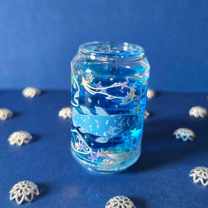 May include: A clear glass jar filled with blue liquid and decorated with a fish and aquatic life design. The jar is surrounded by small, silver, decorative flower-shaped objects on a blue surface.