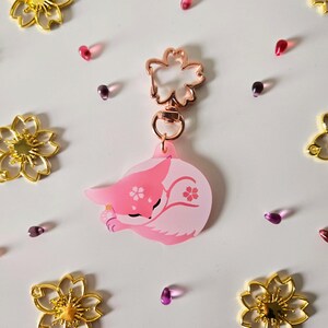 May include: A pink and white fox keychain with a rose gold flower-shaped clasp. The fox is curled up, with cherry blossom details. Gold flower charms and pink beads surround the keychain.