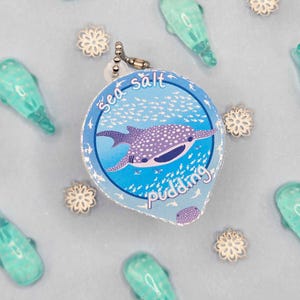 May include: A round, blue and white Sea Salt Pudding charm with a whale shark illustration. The charm is surrounded by small, turquoise whale-shaped charms and silver snowflake-shaped charms on a light blue surface.