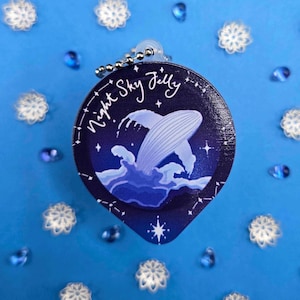May include: A round, dark blue charm with the text "Night Sky Jelly" and a whale breaching the water. The charm is attached to a silver keychain. The background is a light blue surface with snowflake and gem-shaped decorations.
