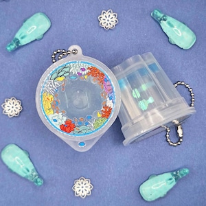 May include: A clear plastic keychain with a colorful coral reef design, open to reveal a small compartment. Surrounding the keychain are small, turquoise-colored dolphin-shaped charms and silver snowflake-shaped embellishments, all on a blue background.