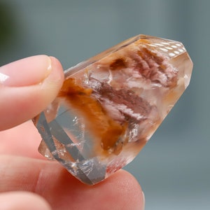 May include: A clear quartz crystal with internal inclusions of reddish-brown and white minerals. The crystal is held between fingers, showcasing its faceted structure and natural beauty. The crystal is a unique specimen.