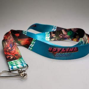 May include: A blue lanyard with a white and green geometric pattern on one side and a colourful graphic of a character from the video game Hotline Miami on the other side. The lanyard has a silver clip.