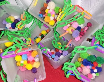 May include: Multiple clear plastic containers with green chain handles filled with colorful pom poms. The containers are arranged on a red surface.