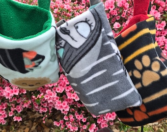 May include: Three hanging fleece pouches with different patterns. The first pouch is light blue with a green lining and a black, white, and orange graphic. The second pouch is gray with white stripes and a black and white graphic. The third pouch is black with orange, yellow, and brown stripes and a brown paw print.