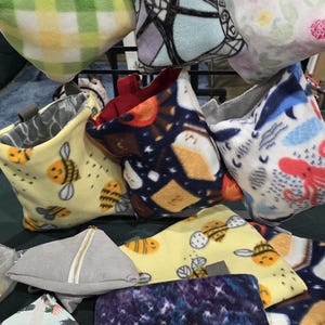 Puede incluir: Una colecci&oacute;n de bolsas de tela de felpa de colores con varios patrones, incluyendo un patr&oacute;n a cuadros, un patr&oacute;n floral, un patr&oacute;n de abejas, un patr&oacute;n de galaxia y un patr&oacute;n con un personaje de dibujos animados. Las bolsas est&aacute;n colgadas en un estante de metal.