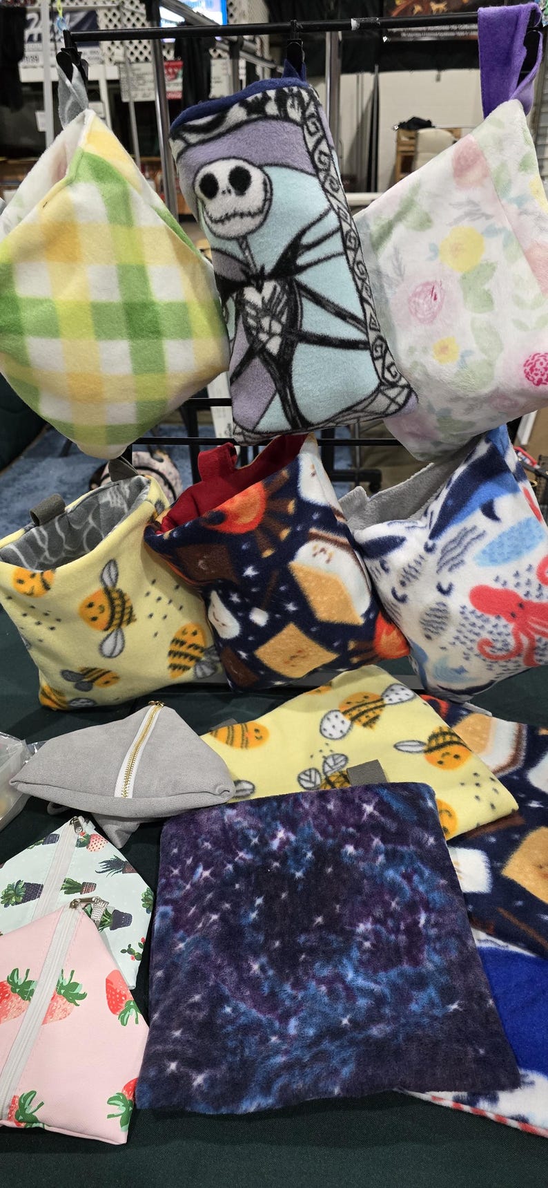 Puede incluir: Una colecci&oacute;n de bolsas de tela de felpa de colores con varios patrones, incluyendo un patr&oacute;n a cuadros, un patr&oacute;n floral, un patr&oacute;n de abejas, un patr&oacute;n de galaxia y un patr&oacute;n con un personaje de dibujos animados. Las bolsas est&aacute;n colgadas en un estante de metal.