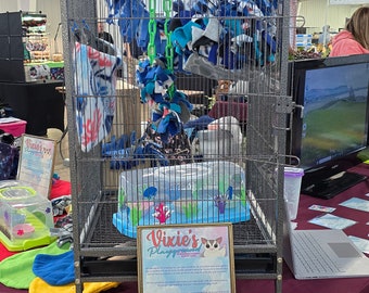 May include: A gray metal cage with a blue and white fleece toy hanging inside. The cage is on wheels and has a sign that says "Vixie's Playground".