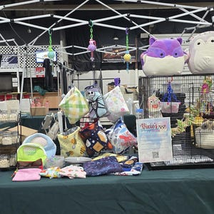 Puede incluir: Un puesto de mesa con diversos art&iacute;culos para mascotas, incluyendo jaulas, juguetes y ropa de cama. La mesa est&aacute; cubierta con un mantel verde. Hay dos jaulas en la imagen, una con un p&aacute;jaro y la otra con un animal peque&ntilde;o. Ambas jaulas son de metal negro con lados de malla de alambre. Los juguetes incluyen una variedad de juguetes de peluche, juguetes de cuerda y juguetes para masticar. La ropa de cama incluye una variedad de materiales, como heno, virutas de madera y papel.
