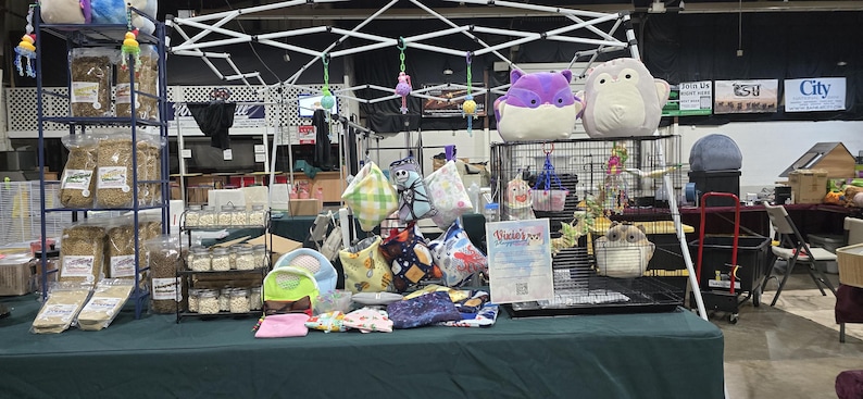Puede incluir: Un puesto de mesa con diversos art&iacute;culos para mascotas, incluyendo jaulas, juguetes y ropa de cama. La mesa est&aacute; cubierta con un mantel verde. Hay dos jaulas en la imagen, una con un p&aacute;jaro y la otra con un animal peque&ntilde;o. Ambas jaulas son de metal negro con lados de malla de alambre. Los juguetes incluyen una variedad de juguetes de peluche, juguetes de cuerda y juguetes para masticar. La ropa de cama incluye una variedad de materiales, como heno, virutas de madera y papel.