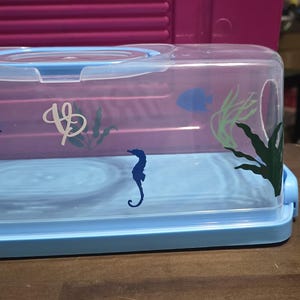 Puede incluir: Un recipiente de plástico transparente con tapa azul y base azul. El recipiente tiene un recorte en el lateral y está decorado con diseños de caballitos de mar y algas.