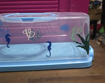 May include: A clear plastic container with a blue lid and a blue base. The container has a cutout on the side and is decorated with seahorse and seaweed designs.
