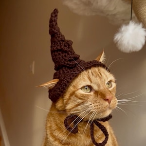 Puede incluir: Un gato atigrado naranja con un sombrero de mago marrón tejido a ganchillo con una punta puntiaguda y una pajarita a juego. El gato está sentado sobre una superficie blanca y esponjosa en forma de nube, con un fondo neutro.