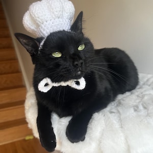May include: A black cat wearing a white crocheted chef's hat and bow tie. The cat has green eyes and is lying on a fluffy white surface. The chef's hat is a traditional style, and the bow tie is a simple design.