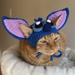 Op de afbeelding: Een rode kat draagt een handgemaakte blauwe en paarse gehaakte hoed met grote oren. De hoed heeft een cartoonachtig ontwerp met grote ogen en een kleine antenne. De kat zit op een zacht, wit oppervlak.