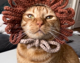 Può includere: Un gatto zenzero che indossa un costume da leone di maglia marrone con un papillon beige. Il gatto guarda direttamente la telecamera con un'espressione seria.