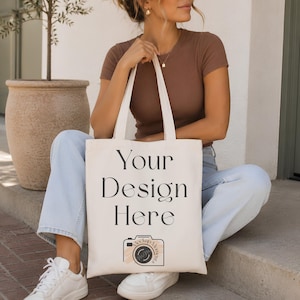 May include: A cream-coloured canvas tote bag featuring the black printed text Your Design Here and a camera icon with the letters MM inside. The bag has two shoulder straps and a rectangular shape, providing a blank space for custom graphic placement.