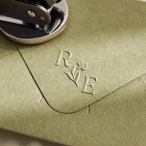 Pode incluir: Uma ferramenta de relevo em metal escuro repousa sobre um envelope de papel verde sálvia. O envelope apresenta um monograma em relevo com as letras R e E separadas por uma pequena ilustração floral na aba do envelope.