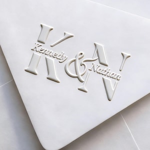 May include: A white envelope featuring a raised, embossed design on the flap. The design includes large letters K and N with an ampersand in the centre. The name Kennedy is printed across the letter K, and the name Nathan is printed across the letter N.