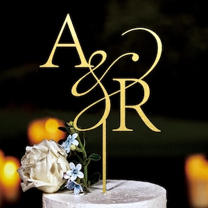 Può includere: Decorazione dorata per torta con le lettere intrecciate "A & R". Il topper è posto su una torta bianca decorata con rose bianche e piccoli fiori blu. Lo sfondo è scuro con luci sfocate.