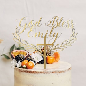 May include: Gold cake topper with the text "God Bless Emily" in a cursive font, surrounded by a wreath of leaves. A gold cross is centred below the text.