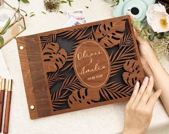 May include: A wooden wedding guest book with a laser-cut tropical leaf design. The cover is engraved with the names "Oliver & Amelia" and the date "08.08.2024". The book is brown with a black background, and is held by a person.