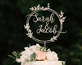 May include: A white wedding cake with a wooden cake topper that says "Sarah & Jacob" in cursive script. The topper is a circle with leaves around it.