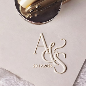 Pode incluir: Um selo de relevo dourado repousa sobre papel rosa claro, com as iniciais em relevo "A&S" e a data "20.12.2026". O selo é parcialmente visível, sugerindo seu uso para criar impressões em relevo. O papel tem uma superfície texturizada.