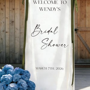 May include: A white fabric banner with the text "WELCOME TO WENDY'S Bridal Shower" and the date "MARCH 7TH, 2026". The banner is adorned with green ribbons and a basket of blue flowers.
